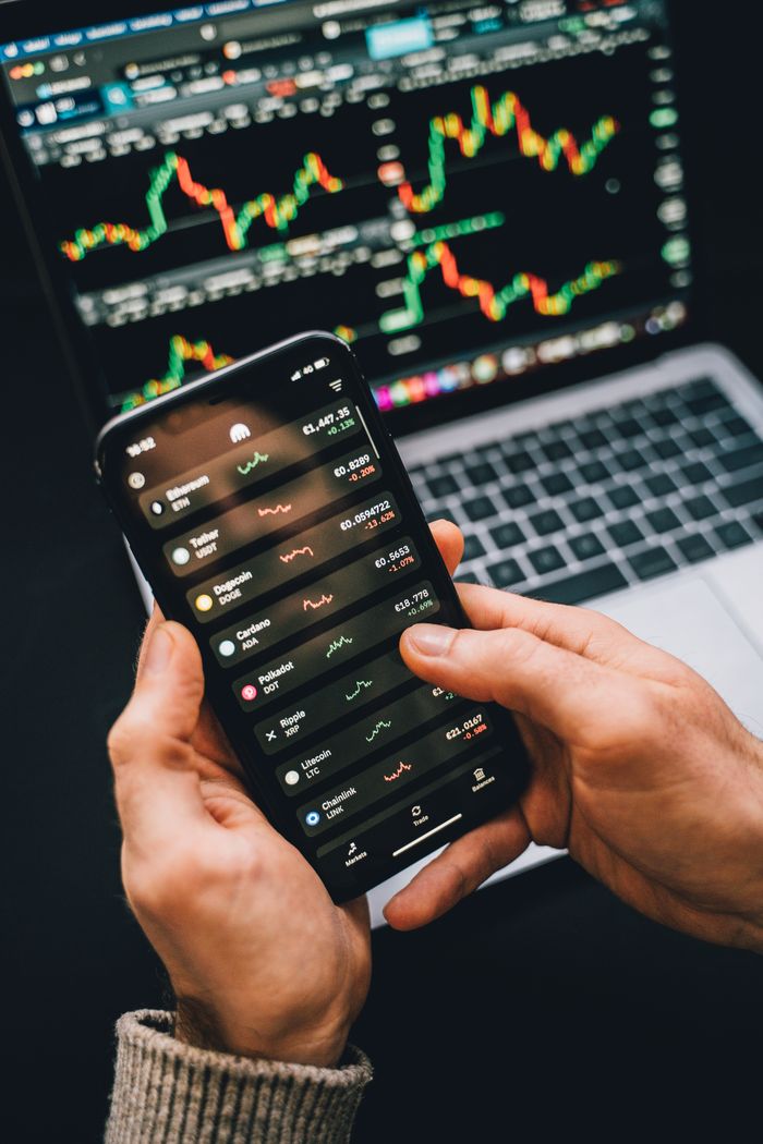 about-us-02 Des mains tiennent un smartphone avec une application boursière, tandis qu’un ordinateur portable affiche des graphiques. Idéal pour les univers de la finance et de la technologie.